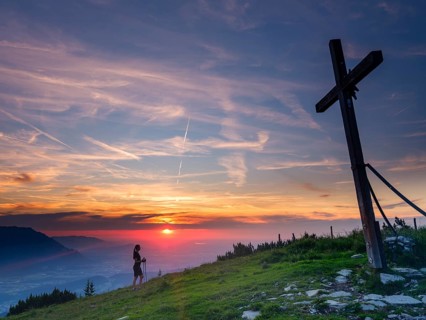Young slim woman with hiking poles next to a summit cross at sunset, cirrus clouds in the sky,