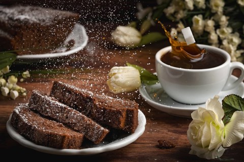 A cup of coffee, cake with icing sugar and splashing coffee drops, flower arrangement on a wooden