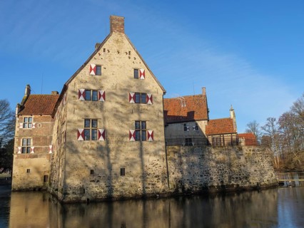A historic moated castle with several windows and red shutters, on the banks of a pond,