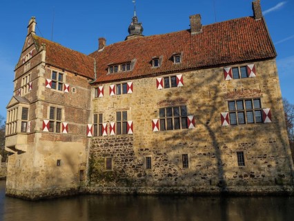 A moated castle with red and white shutters and a red roof, surrounded by water, lüdinghausen,