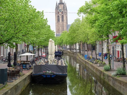 A calm river with boats and green trees on both sides, with a church tower in the background,