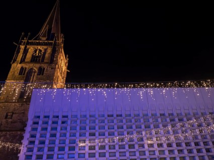 An imposing church tower and a modern building side by side, with festive lighting at night, Ahaus,