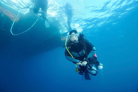 A diver with a camera swims underwater next to a boat on the surface. Dive site Marine reserve Cap