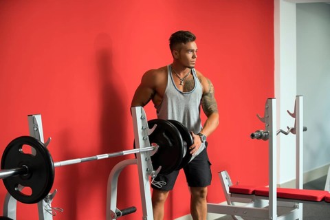 In gym. Tattooed young athlete standing near barbell