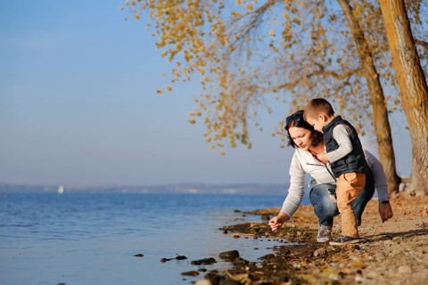 A woman and a child are by a lake on an autumn day, picking something up from the shore while trees