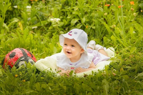 A smiling baby laying on the grass with a white hat, surrounded by flowers and greenery in the