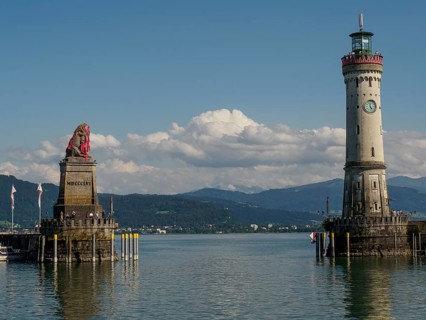 Lion statue and lighthouse with clear sky and calm water, nearby town and mountains in the