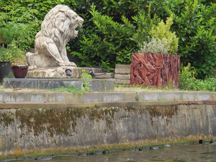 Stone statue of a lion on an overgrown and watered stone bank surrounded by nature, Leiden,