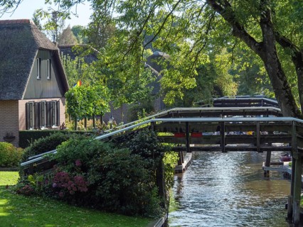 Wooden bridge over a canal, surrounded by trees and a house, with shadow effect through the leaves,