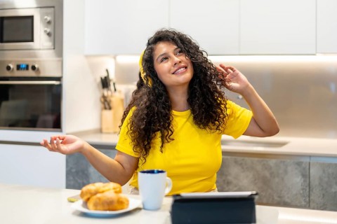 Latin happy young woman dancing while listening to music in the morning in the kitchen at home