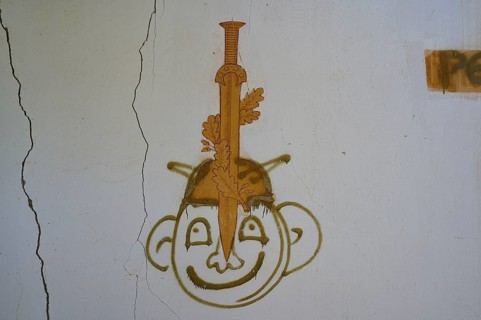 Graffiti of a face with a sword can be seen on a cracked white wall of an abandoned building, Lost