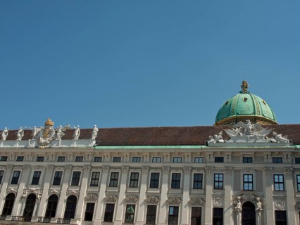 White baroque building with green domed roof and statues on the long façade under a blue sky,