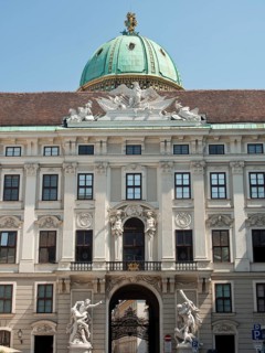 Historic baroque building with green domed roof, archway and statues under a clear sky, Vienna,