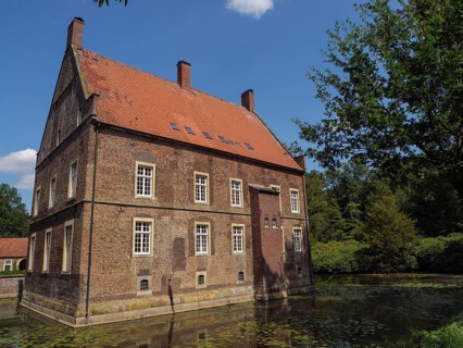 Old brick house with red roof on the edge of a pond, surrounded by green trees in summer, ochtrup,