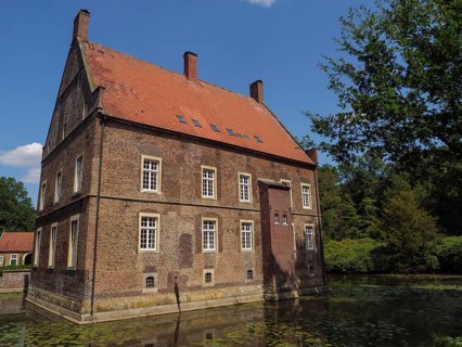 Large brick building by a pond with calm water, surrounded by trees and sunny weather, ochtrup,