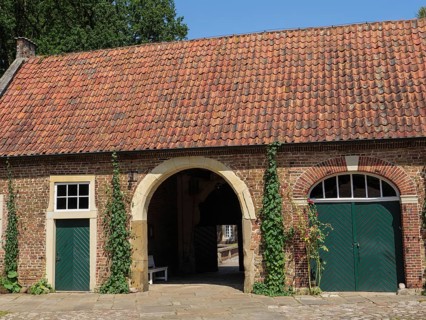 A brick house with a round arch, red tiles on the roof, green doors and surrounded by climbing