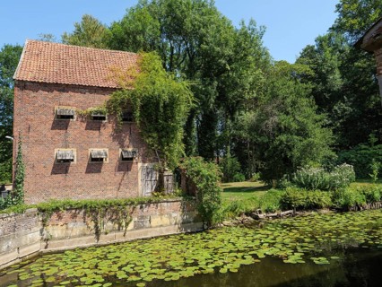 Red brick building with wooden shutters and ivy growth by the pond, surrounded by trees and sunny