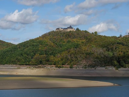 A striking castle overlooks the scene from a wooded hill sloping down to the banks of a river,