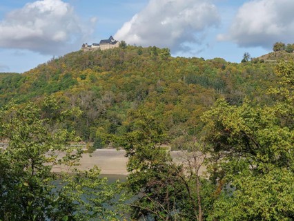 A majestic castle complex stands on a wooded hill that slopes down to a wide, river-like body of
