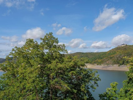 View of the lake and surrounding wooded hills with a castle in the sunny, slightly cloudy sky,