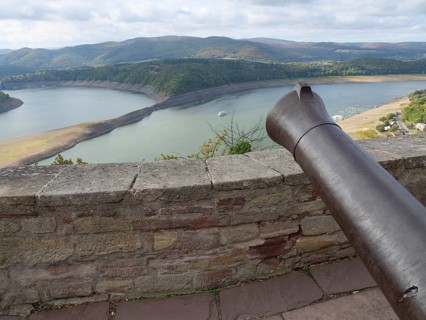 View from a stone wall with a cannon onto a lake surrounded by wooded hills under a cloudy sky,