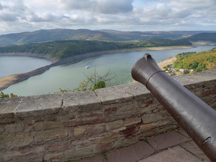 View from a stone wall with a cannon onto a lake surrounded by wooded hills under a cloudy sky,
