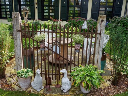 A rustic garden gate with statues and various plants surrounded by green landscape in front of a