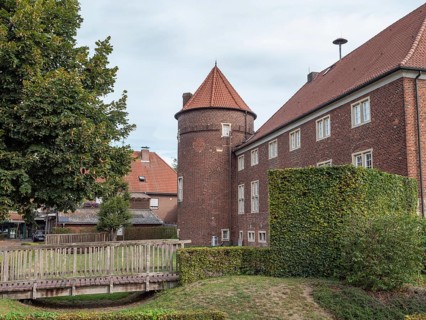 Brick building with tower and roof, surrounded by a hedge and trees, with a small bridge in the