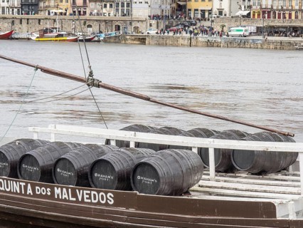 A boat loads wine barrels on the bank of a river while passers-by walk by in the background, Porto,