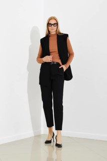 Serious woman in black vest, terracotta turtleneck, and breeches standing against a white office