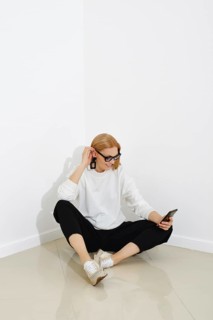 Cheerful woman in sweater and trousers sits on a floor with smartphone in her hand