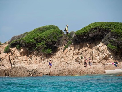 People walking along a rocky coastline, overgrown cliffs in the background and blue sea, ajaccio,