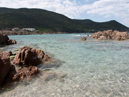 A rocky coastal landscape with clear water and green mountains in the background under a cloudy