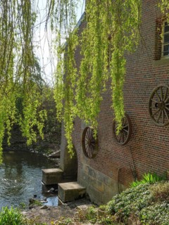 A brick building with large wooden wheels sits on the riverbank, surrounded by green trees and