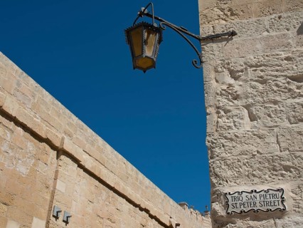 A decorative lamppost and street sign along an old stone wall under a blue sky, mdina,