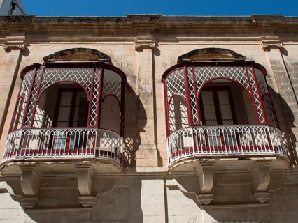 Two ornate and decorative iron balconies in front of a historic stone wall with windows, mdina,