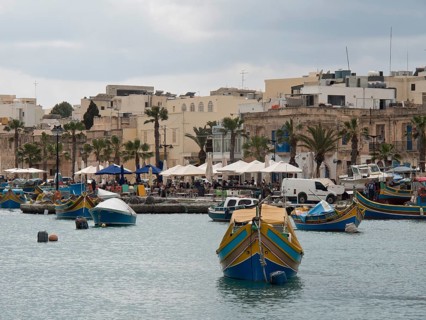 A row of fishing boats in front of a small town with palm trees, under a cloudy sky, marsaxlokk,