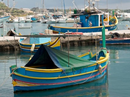 A blue and yellow fishing boat lies calmly in the water of a harbour, marsaxlokk, mediterranean