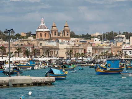 Boats in the harbour with an urban backdrop and a cathedral under a cloudy sky, marsaxlokk,