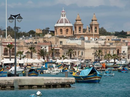 Fishing boats in the harbour with a cathedral in the background under a cloudy sky, marsaxlokk,