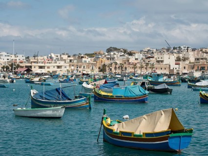 A harbour full of fishing boats with buildings in the background under a cloudy sky, marsaxlokk,