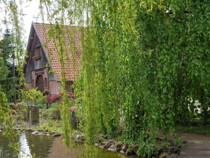A half-timbered house with a tiled roof, surrounded by a willow and a pond, with blooming flowers