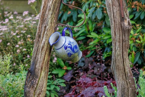 A decorative blue and white jug hangs from a rustic wooden frame in a flowery garden, Borken,