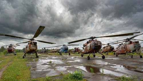 Several old, rusty helicopters are standing on an abandoned and overgrown area under an overcast