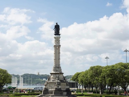 Marble monument with statue on a pedestal, surrounded by green areas and trees under a blue sky