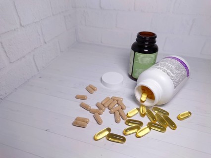 Two open bottles spilling various pills and capsules onto a white table against a white brick wall