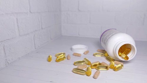 An open bottle of vitamin capsules has spilled yellow and beige pills onto a white table against a