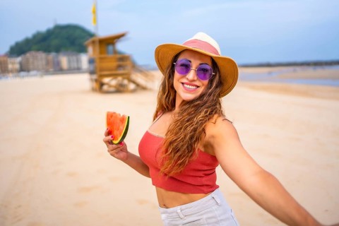 A woman is smiling and holding a watermelon slice. She is wearing a straw hat and sunglasses