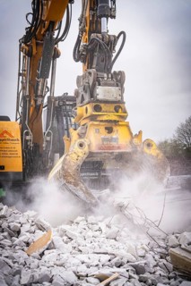 Large yellow excavator with grab crushes concrete rubble and stirs up dust on a construction site,