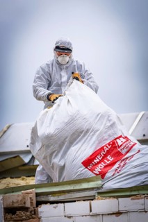 Worker in protective suit and mask handling a large bag of fibre wool on a construction site,